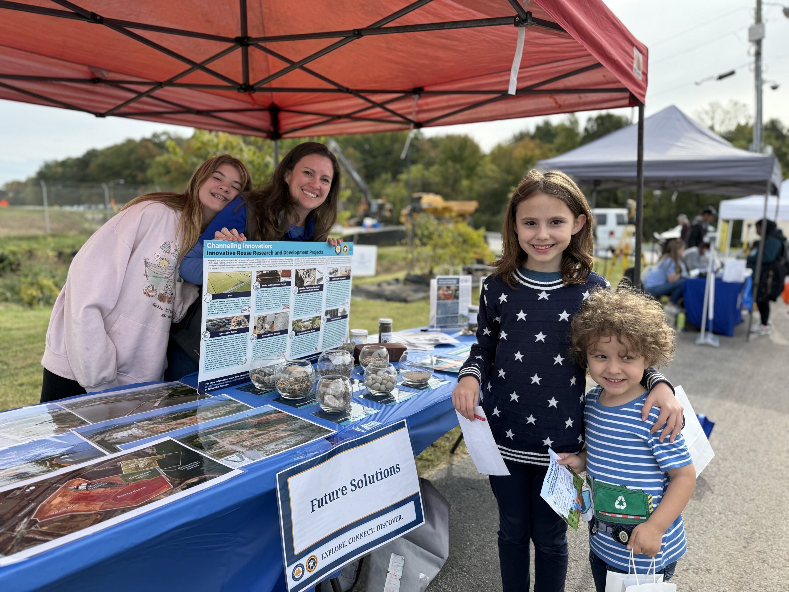 Two women are presenting a Future Solutions booth and discussing information with a young girl and boy at the 2025 Cox Creek Open House, now called Port of Baltimore Open House.
